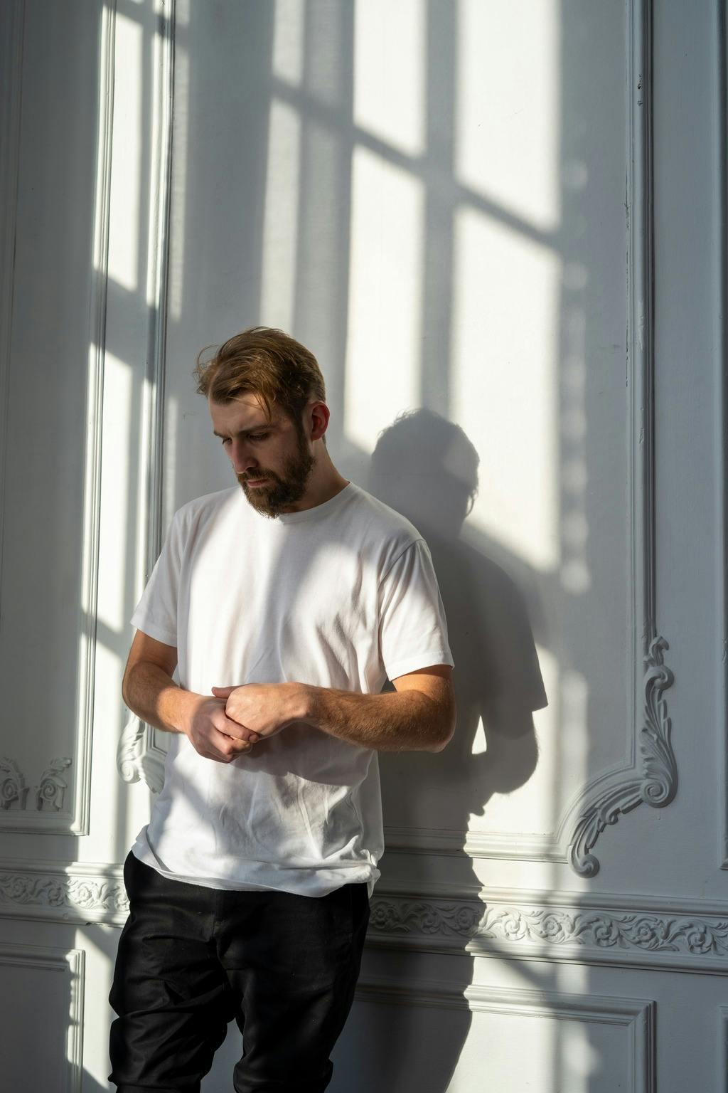 Man in t-shirt leans against a wall and looks down forlornly.