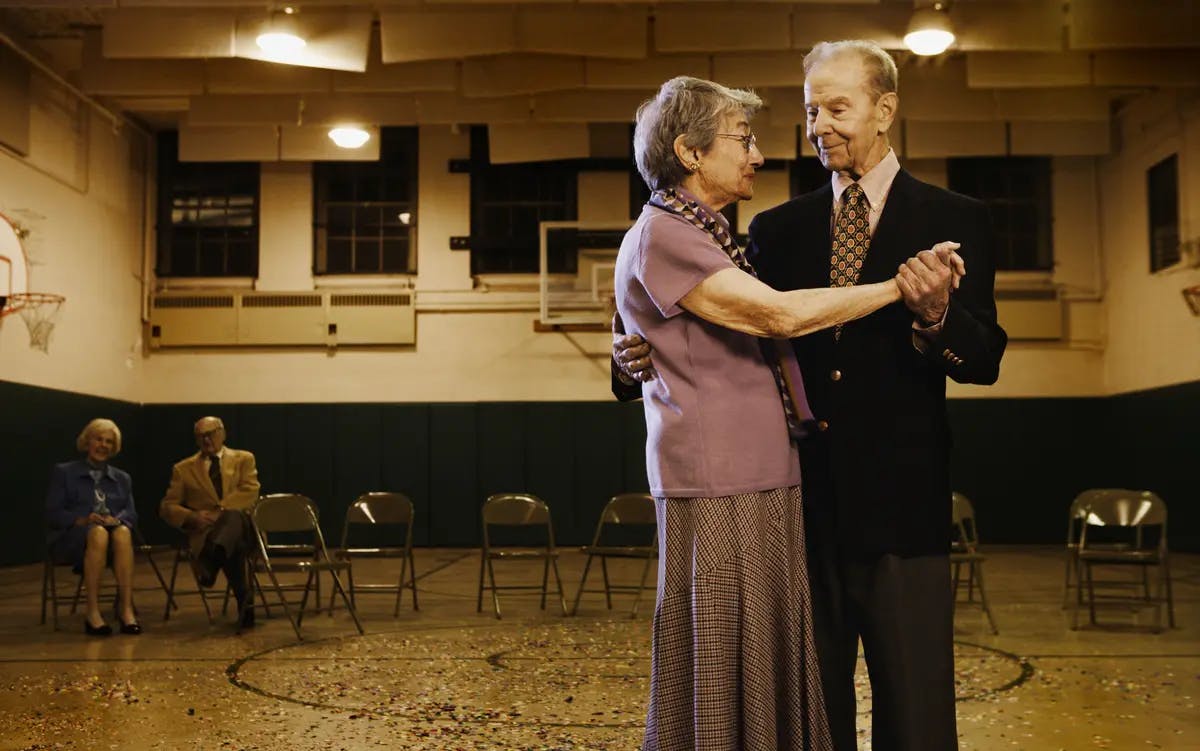 Older couple in black tie dress dance alone in the foreground while another couple looks on from the background.