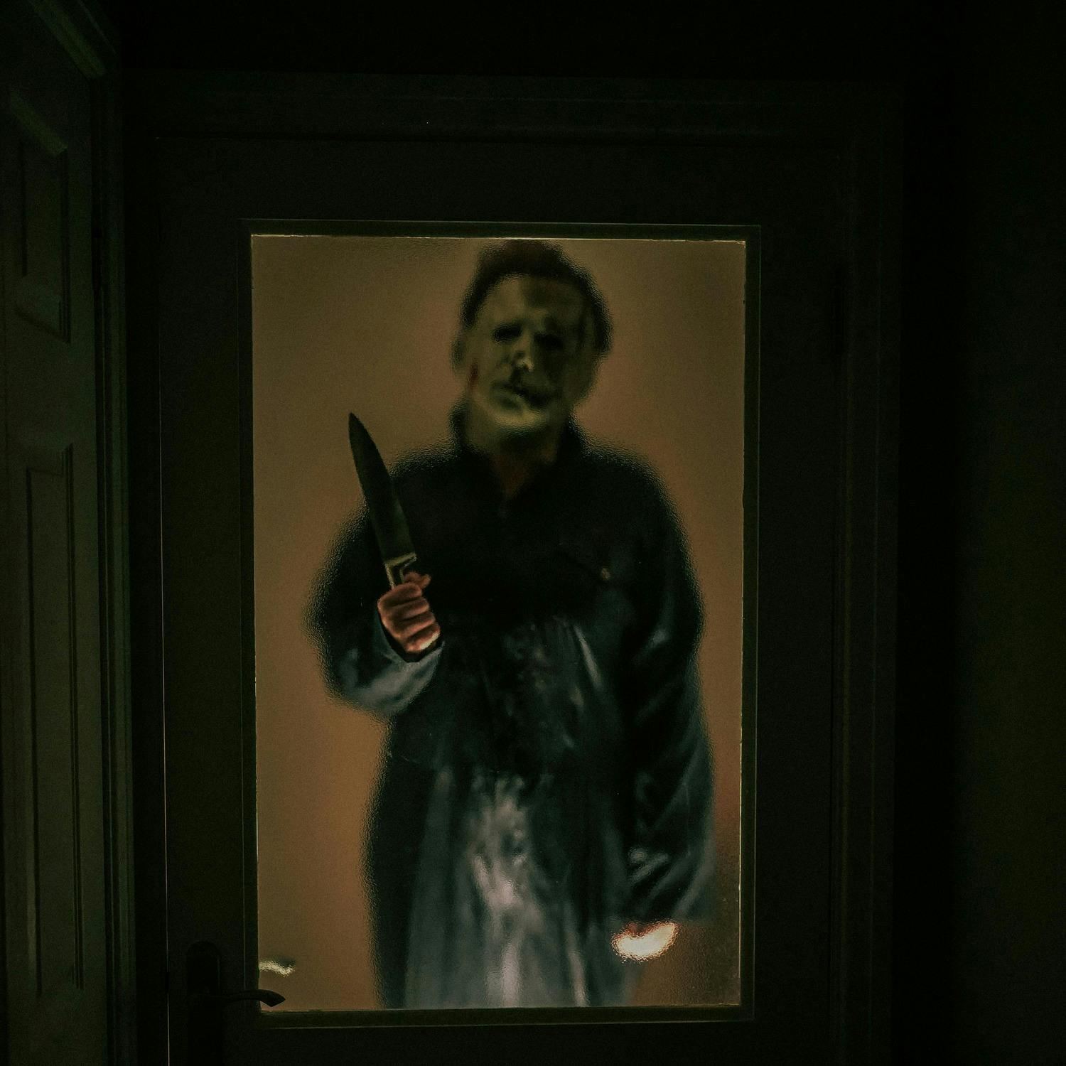Photo of person holding a bloody knife looking through a glass door.