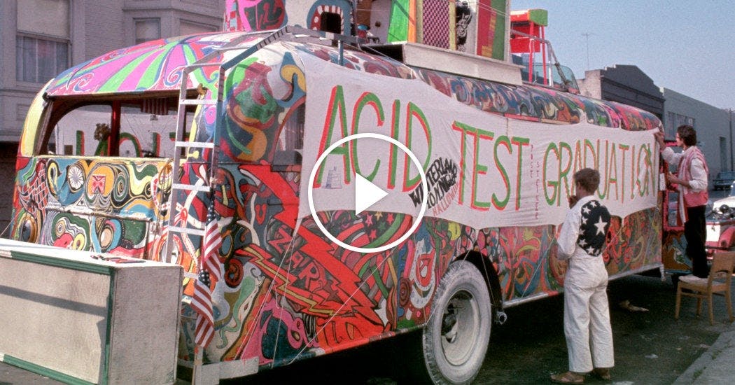 A long sign that reads, "acid test graduation" hangs off of a bus painted with intersecting and overlapping colors and shapes. 