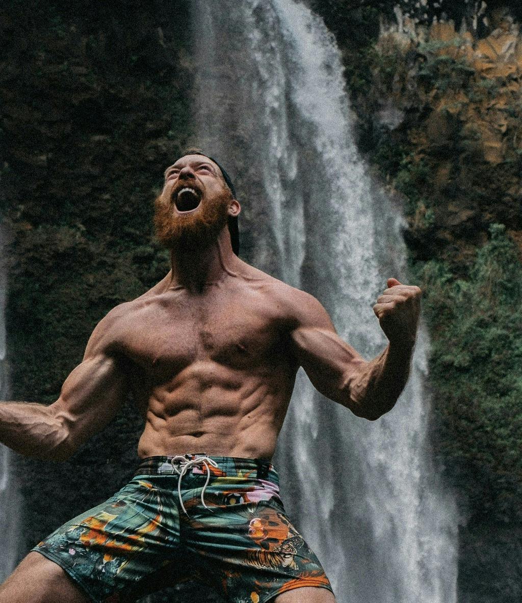 Photo of person screaming toward the heavens as they flex their muscles before a waterfall.