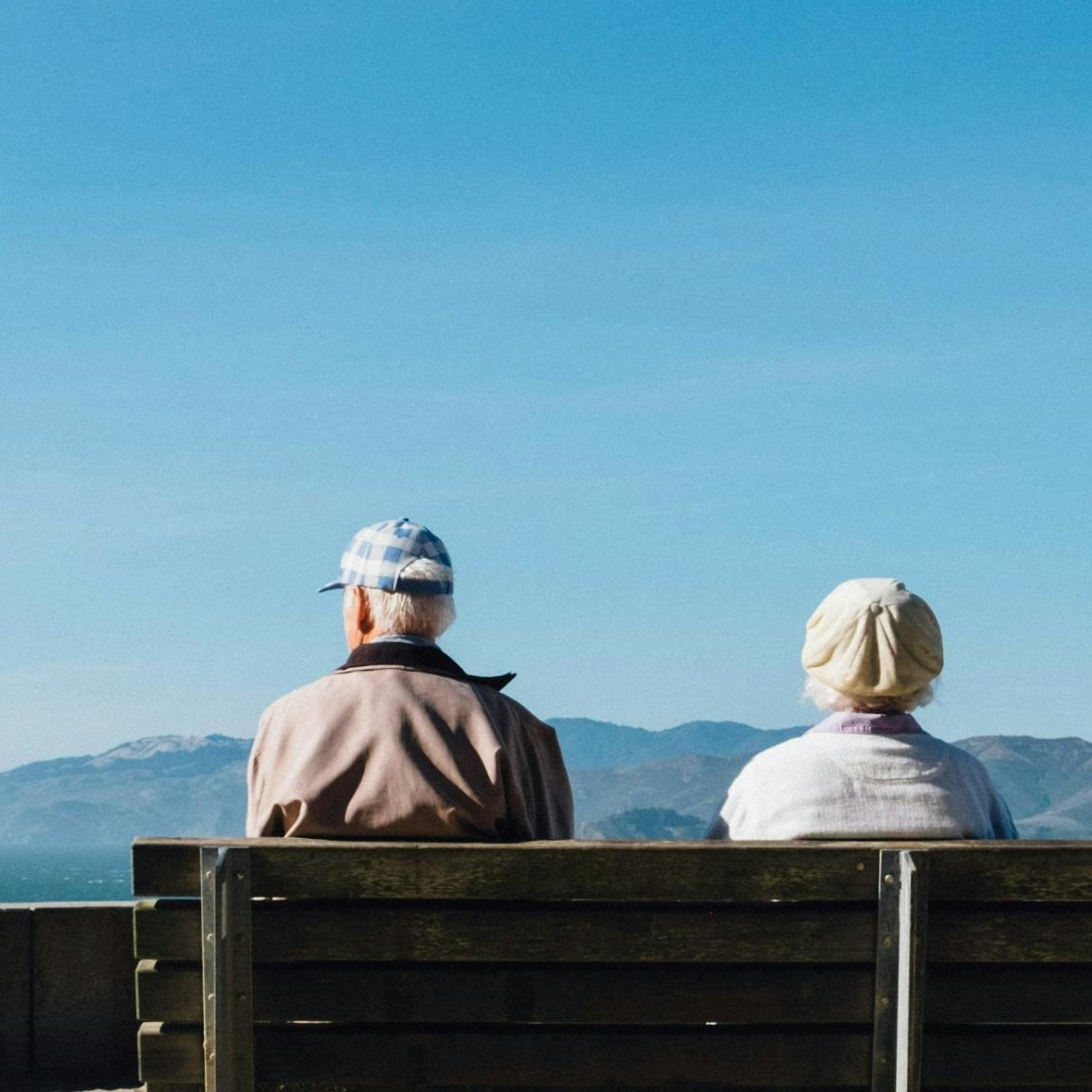 Photo of two older people sitting on a bench facing away from the camera.