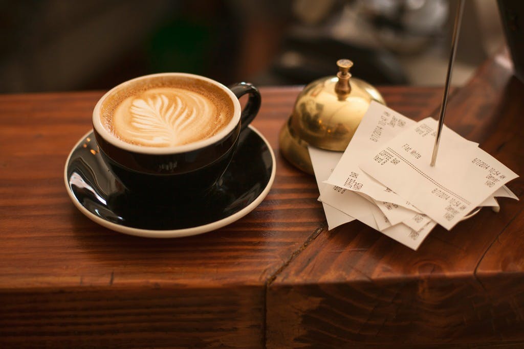 A coffee cup and saucer sit on a bar to the left of a service bell and impaled stack of receipts.